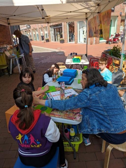 Foto einer Straßenszene bei gutem Wetter: An improvisierten Tischen sitzen Kinder und malen etwas zusammen mit einer erwachsenen Person.