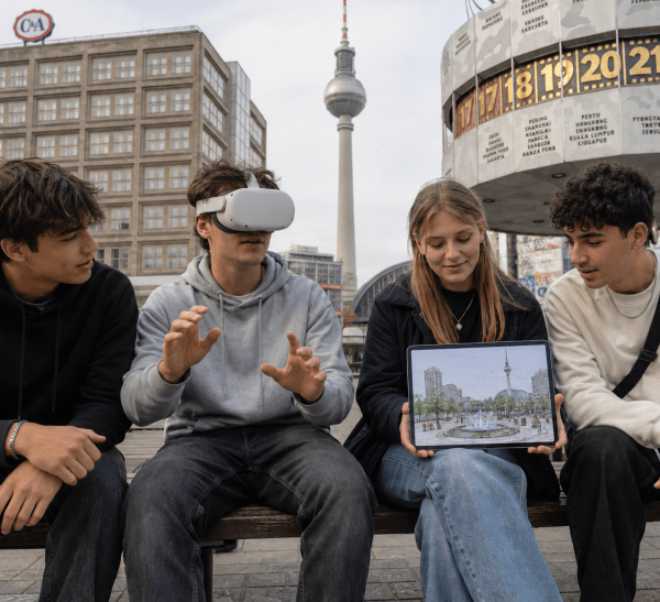Vier Jugendliche mit Laptop und VR-Brille, die draußen sitzen, im Hintergrund der Berliner Fernsehturm und die Weltzeituhr am Alexanderplatz